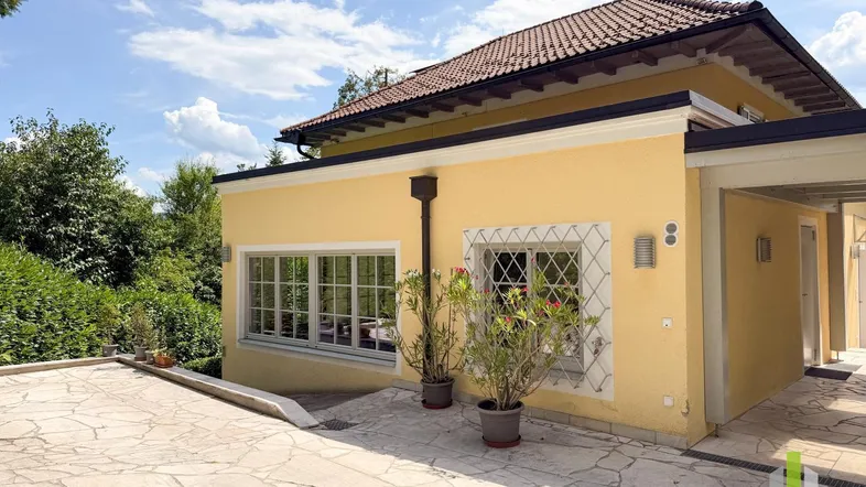 Gelbe Hausfassade mit gepflastertem Vorplatz und grüner Umgebung unter blauem Himmel.