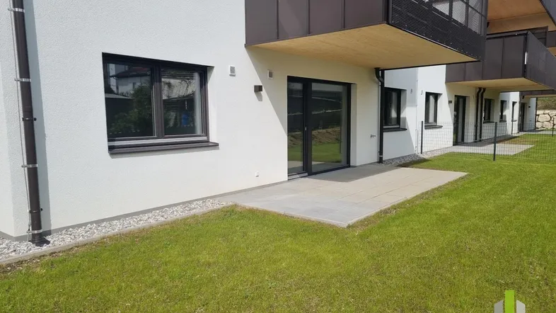 Außenansicht eines Neubaus mit kleiner gepflasterter Terrasse und Rasenfläche, darüber ein Balkon.