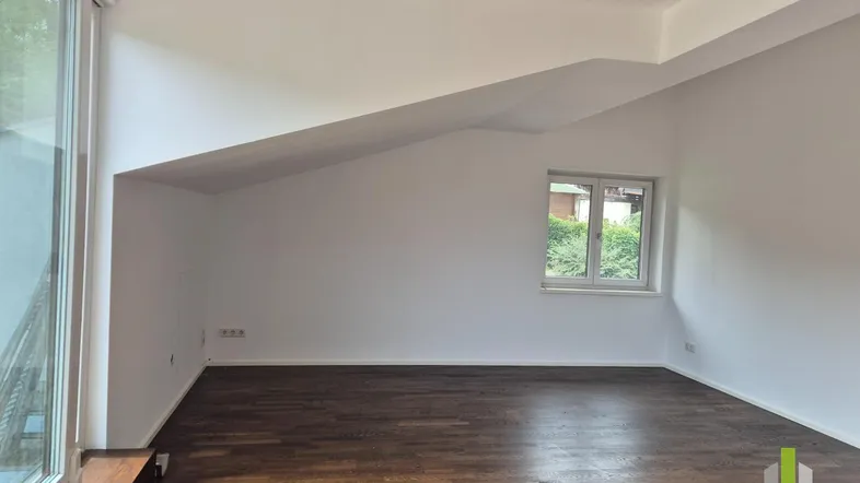 Helles Zimmer mit Dachschräge, dunklem Holzboden und Zugang zu einem Balkon oder einer Terrasse.