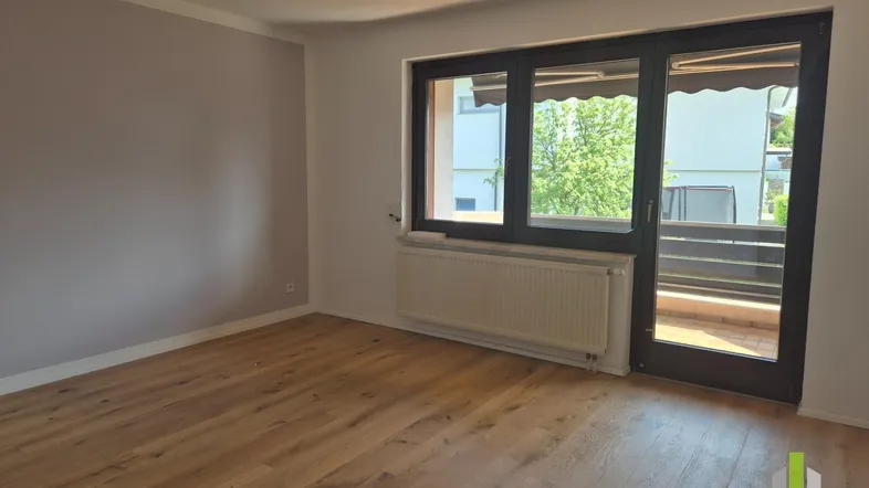 Helles Zimmer mit Holzboden, einer Akzentwand und Zugang zu einem Balkon mit Blick ins Grüne.