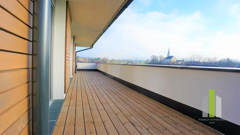 Geräumige Dachterrasse mit Holzboden und weitem Blick auf die umliegende Landschaft und eine Kirche.