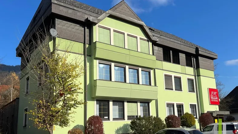 Grünes Gebäude mit mehreren Fenstern und einem dunklen Dach unter blauem Himmel.