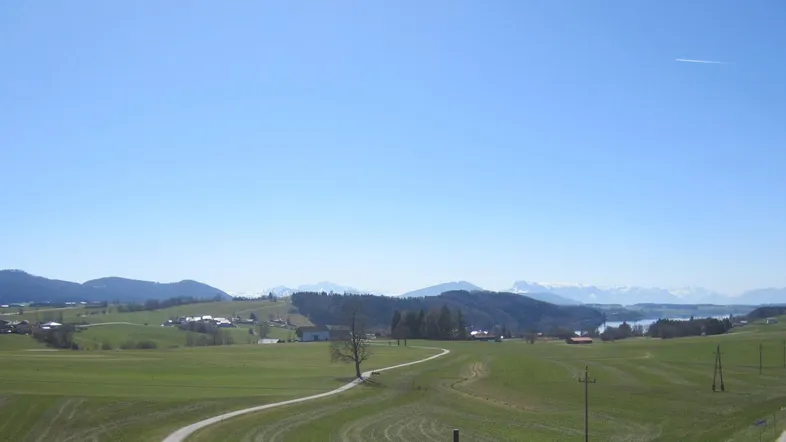 Weitläufiger Blick über grüne Hügel und Felder mit Bergen und einem See in der Ferne.