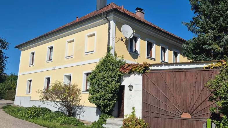 Gelbes Landhaus mit rotem Ziegeldach und Holztor, umgeben von grüner Vegetation unter blauem Himmel.