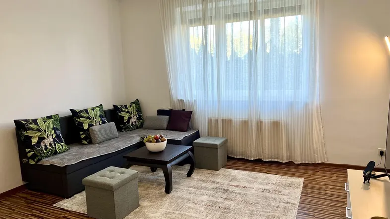 Gemütliches Wohnzimmer mit Ecksofa, Couchtisch und großem Fenster mit hellen Vorhängen.