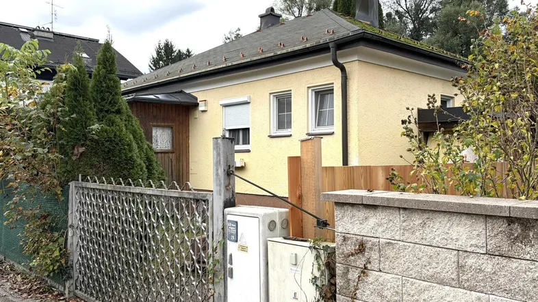 Gelbes Einfamilienhaus mit Zaun und Bäumen, umgeben von Vegetation unter bewölktem Himmel.