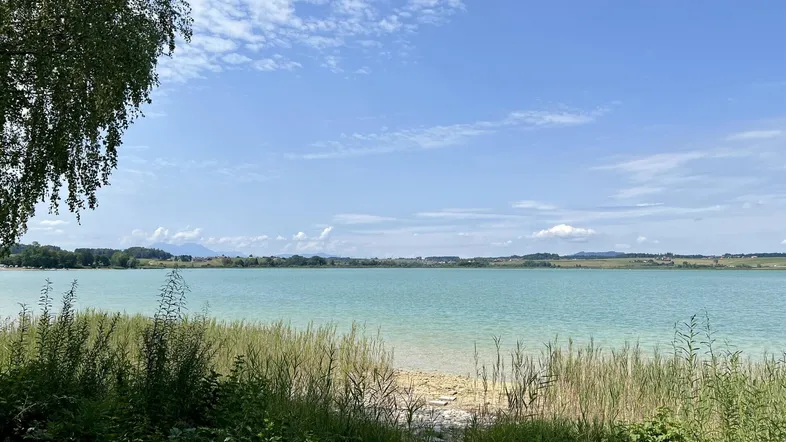 Idyllischer Blick auf einen ruhigen See mit grünen Ufern und blauem Himmel an einem sonnigen Tag.