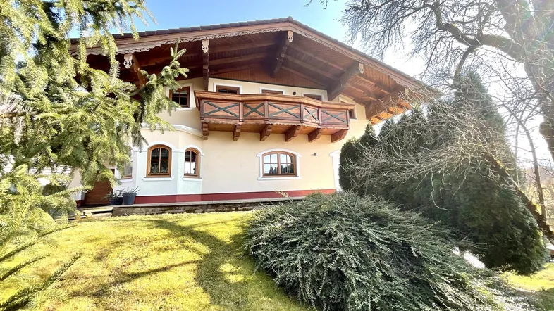 Traditionelles Haus im alpenländischen Stil mit Holzbalkon und gepflegtem Gartenbereich unter strahlender Sonne.