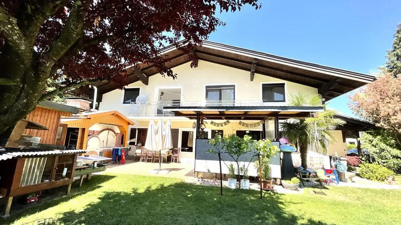 Haus mit großem Garten, Terrasse und Außenküche unter einem Baum an einem sonnigen Tag.