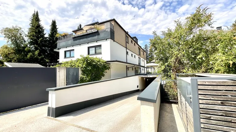 Modernes Mehrfamilienhaus mit weißer Fassade, Balkonen, Dachterrasse und Zufahrt zur Tiefgarage.