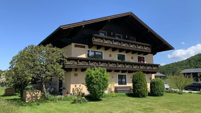 Traditionelles Haus im Alpenstil mit Holzbalkonen und gepflegtem Gartenbereich unter blauem Himmel.