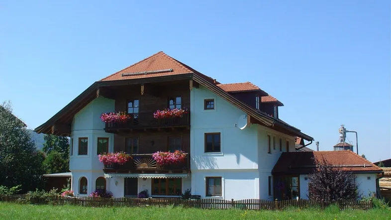 Traditionelles Haus mit blühenden Balkonen und rotem Ziegeldach, umgeben von grünen Wiesen.