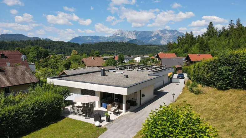 Modernes Haus mit Flachdach, Solaranlage und großer Terrasse, umgeben von grüner Landschaft und Bergen.