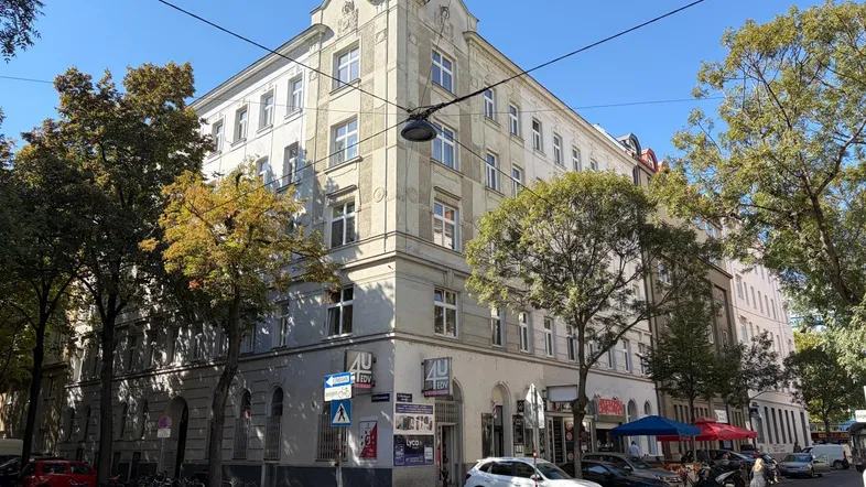 Historisches Eckgebäude mit klassischer Fassade und Bäumen an einer belebten Straße unter blauem Himmel.