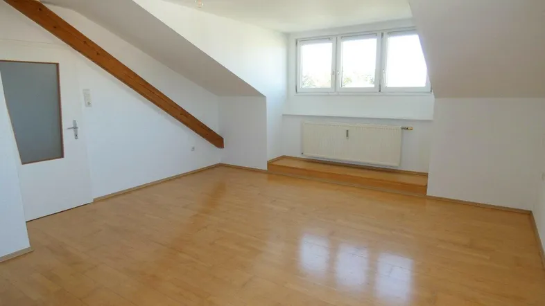 Helles Zimmer im Dachgeschoss mit Holzfußboden, schrägen Wänden und einem Fensterbereich.