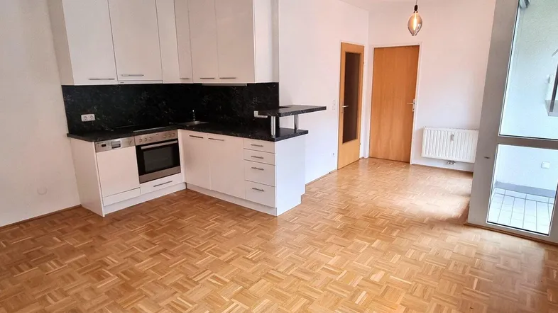 Heller offener Wohnbereich mit integrierter Küchenzeile, Parkettboden und Zugang zum Balkon.