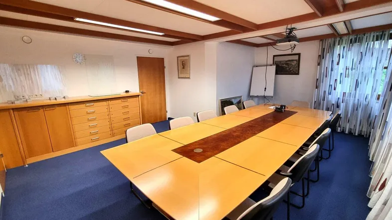 Geräumiger Konferenzraum mit großem Holztisch, blauer Teppichboden und sichtbaren Deckenbalken, ideal für Besprechungen.