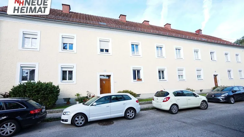 Mehrfamilienhaus mit heller Fassade, Rollläden an den Fenstern und parkenden Autos auf der Straße.