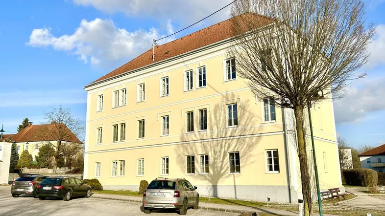 Gelbes Mehrfamilienhaus mit rotem Dach und Parkplätzen unter blauem Himmel.