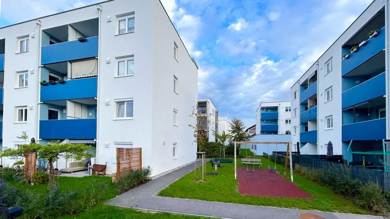 Mehrere moderne Wohngebäude mit blauen Balkonen und einem Spielplatz im Grünen.