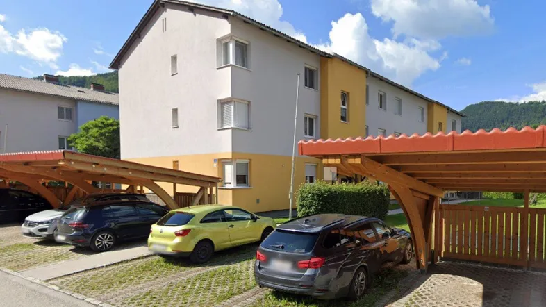 Mehrfamilienhaus mit gelb-weißer Fassade, umgeben von Grünflächen und Carports, mit Bergblick im Hintergrund.