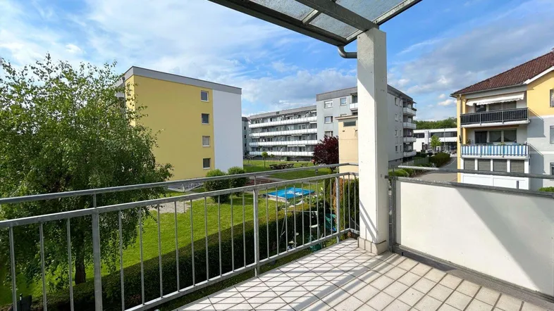 Geräumiger Balkon mit Fliesenboden und Überdachung, bietet Blick auf die umliegende Grünfläche und Wohngebäude.