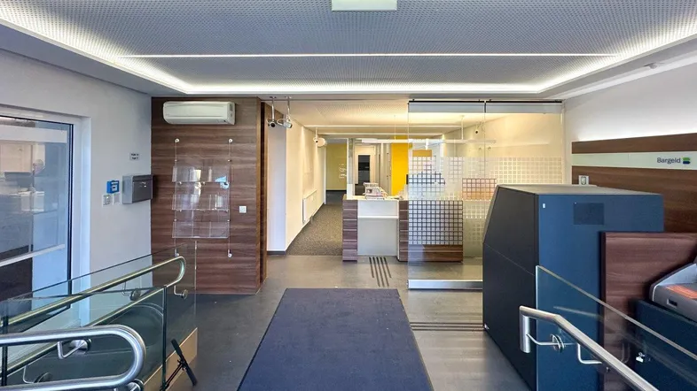 Geräumiger Eingangsbereich mit Rampe, Geldautomaten und modernen Holzverkleidungen unter perforierter Decke.