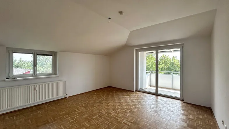 Helles Zimmer mit Parkettboden, Dachschräge und Zugang zu einem kleinen Balkon.