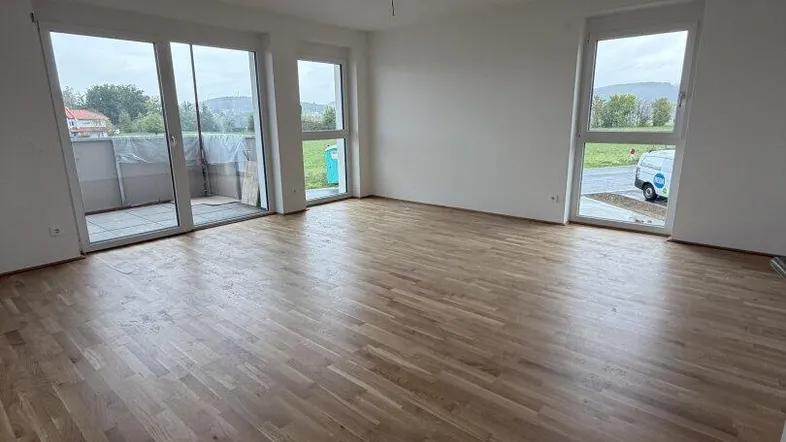 Helles, leeres Wohnzimmer mit Holzboden und Zugang zu einem Balkon. Große Fenster lassen viel Tageslicht herein.