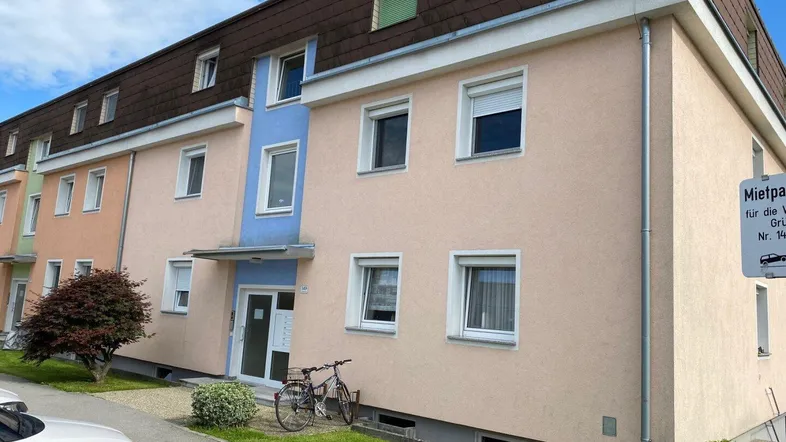 Mehrfamilienhaus mit mehreren Etagen, Fenstern und einem Eingangsbereich, umgeben von Grünflächen und Parkplätzen.
