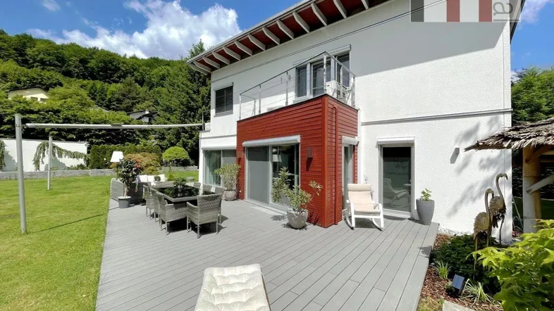 Große Holzterrasse mit Esstisch und Liegestühlen vor dem modernen Haus mit roter Akzentfassade.