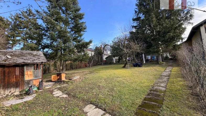 Großer Garten mit Rasenfläche, alten Bäumen und einem kleinen Holzschuppen unter blauem Himmel.