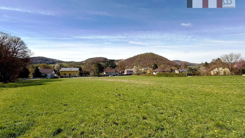 Weitläufiges grünes Feld mit Häusern und Hügeln im Hintergrund unter blauem Himmel.