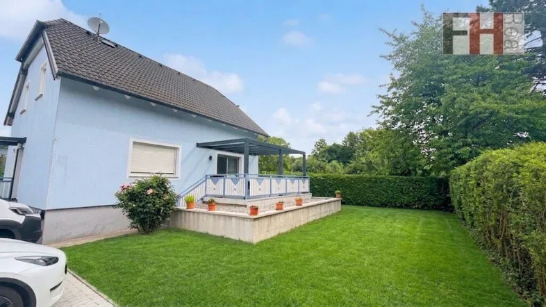 Blaues Einfamilienhaus mit gepflegtem Rasen, Terrasse und Hecke im Gartenbereich.