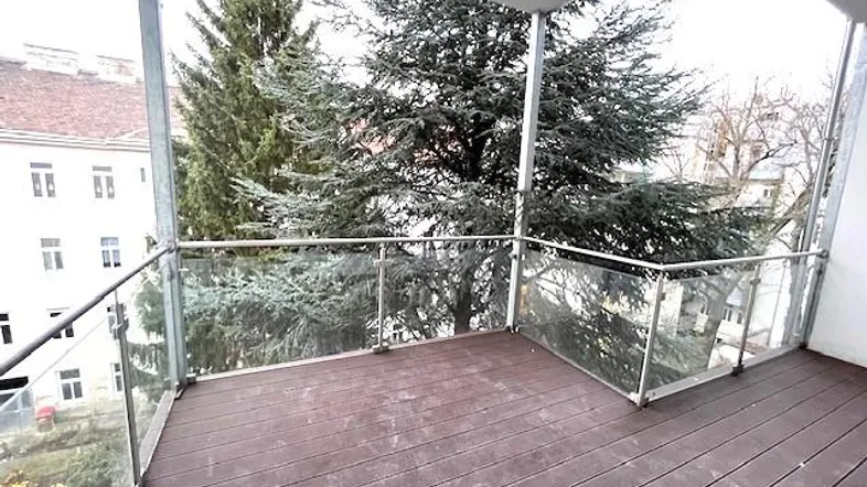 Geräumiger Balkon mit Holzboden und Glasgeländer, umgeben von Bäumen und Blick auf Gebäude.