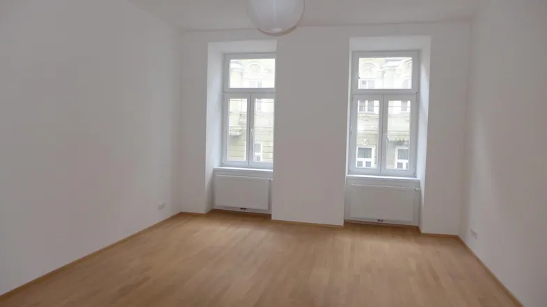 Helles, leeres Zimmer mit Parkettboden und zwei Fenstern, bereit zur individuellen Gestaltung.
