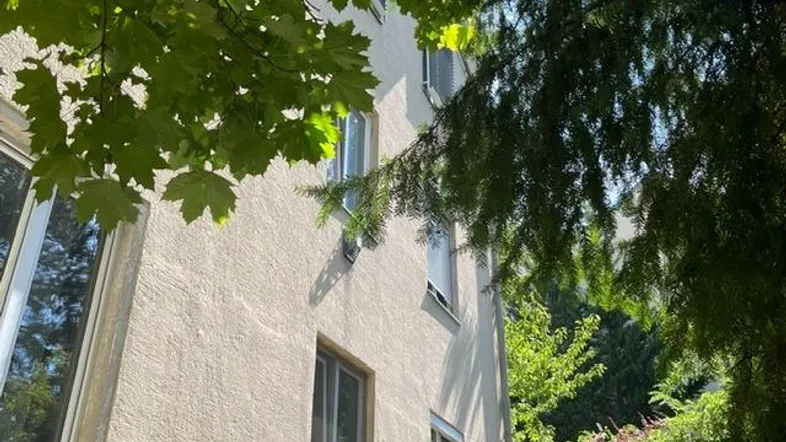 Fassade eines Wohngebäudes, umgeben von grünen Bäumen und Sträuchern, bei Sonnenschein.