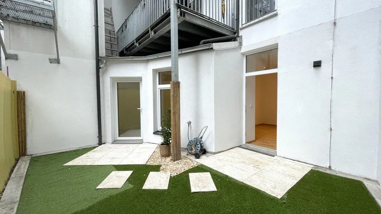Kleiner, pflegeleichter Gartenbereich mit Kunstrasen und Steinplatten, umgeben von weißen Wänden.