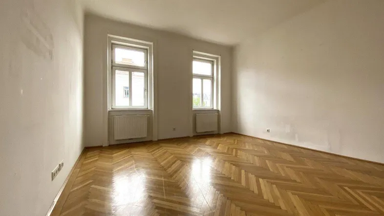 Helles Zimmer mit Fischgrätparkettboden, weißen Wänden und zwei Fenstern, die Tageslicht hereinlassen.