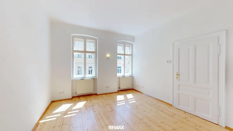 Helles Wohnzimmer mit Holzdielenboden, hohen Decken und zwei großen Fenstern, die viel Tageslicht hereinlassen.