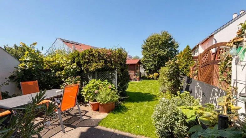 Sonniger Außenbereich mit gepflasterter Terrasse, Gartenmöbeln und Blick auf den gepflegten Rasen und Garten.
