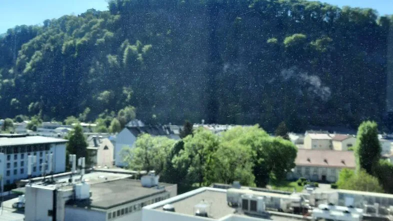 Weitblick auf die umliegende Stadtlandschaft und bewaldete Berge durch ein großes Fenster.