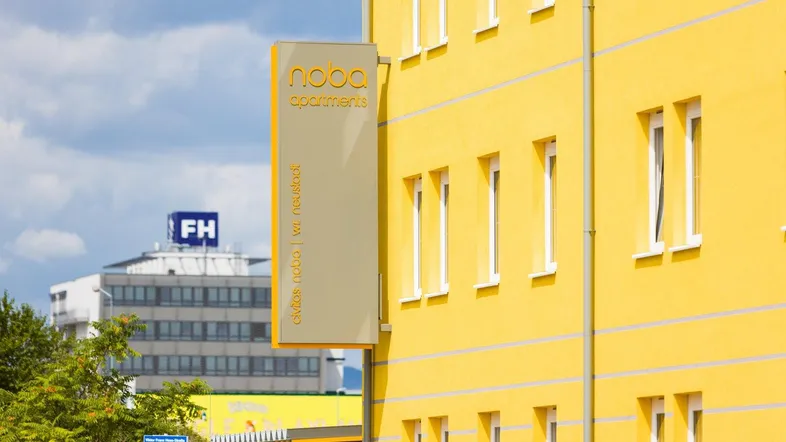 Gelbes Gebäude mit einem Schild für "noba apartments" und einem weiteren Gebäude im Hintergrund.