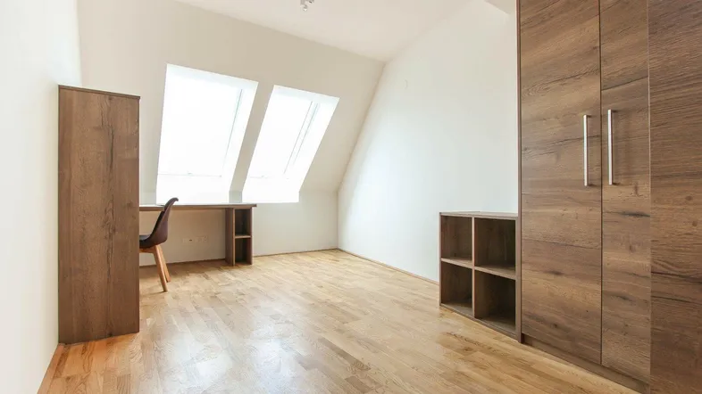 Helles Dachzimmer mit Holzboden, Schreibtisch und Einbauschränken unter der Dachschräge.