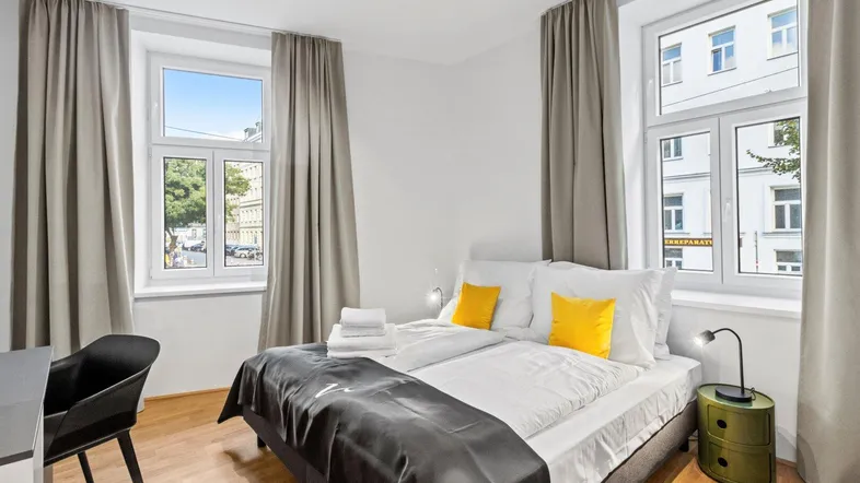 Helles Schlafzimmer mit Doppelbett, Holzboden und Blick auf die Straße durch große Fenster.