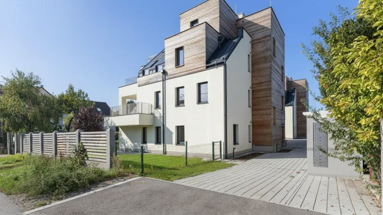 Moderne Wohnanlage mit Holz- und Putzfassade, umgeben von Grünflächen und Zufahrtswegen unter blauem Himmel.