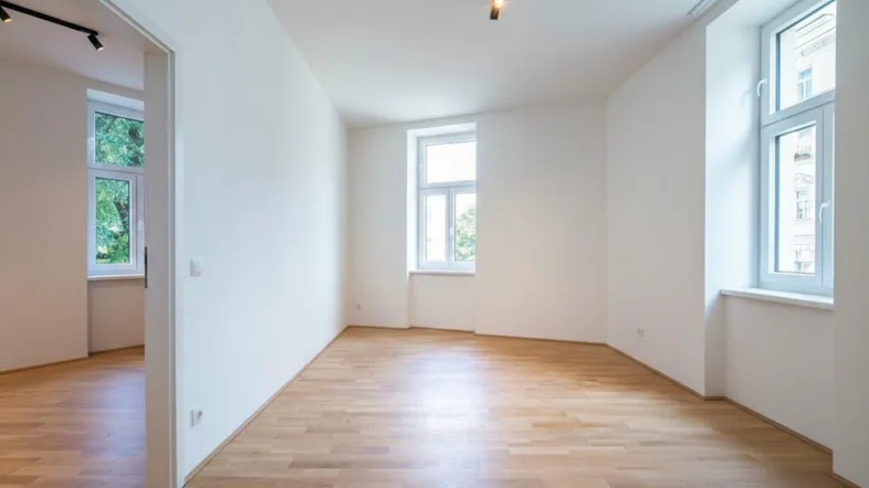 Helles, leeres Zimmer mit Holzboden und zwei großen Fenstern, bereit zur individuellen Gestaltung.