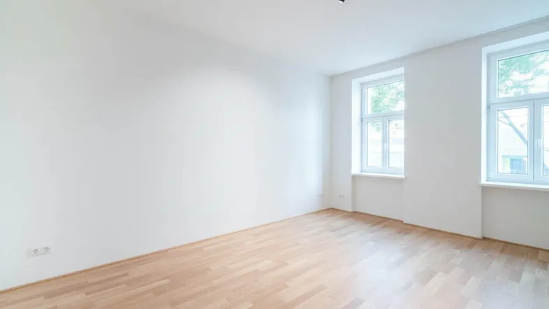 Heller, leerer Wohnraum mit hohen Decken, großen Fenstern und neuem Holzfußboden.