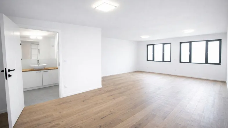 Leerer, heller Wohnraum mit Holzboden und Zugang zu einem modernen Badezimmer. Große Fenster lassen viel Tageslicht herein.
