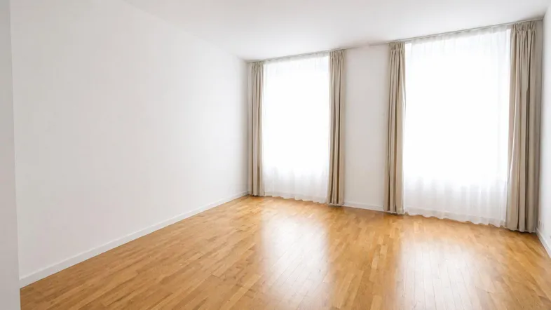 Heller Raum mit weiß gestrichenen Wänden, Holzparkettboden und zwei großen Fenstern mit Vorhängen.